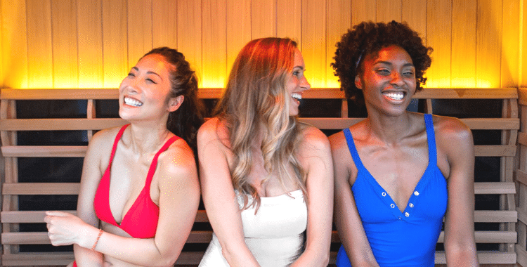 A group of 3 women in swimsuits sitting and laughing in a sauna