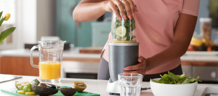 Woman standing at a kitchen counter with chopped veggies & fruit, blending them in a food processor