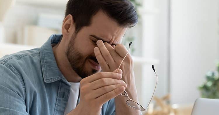 Man holding his glasses in one hand and pinching the bridge of his nose with the other hand as his brow furrows.