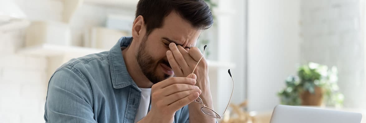 Man holding his glasses in one hand and pinching the bridge of his nose with the other hand as his brow furrows.