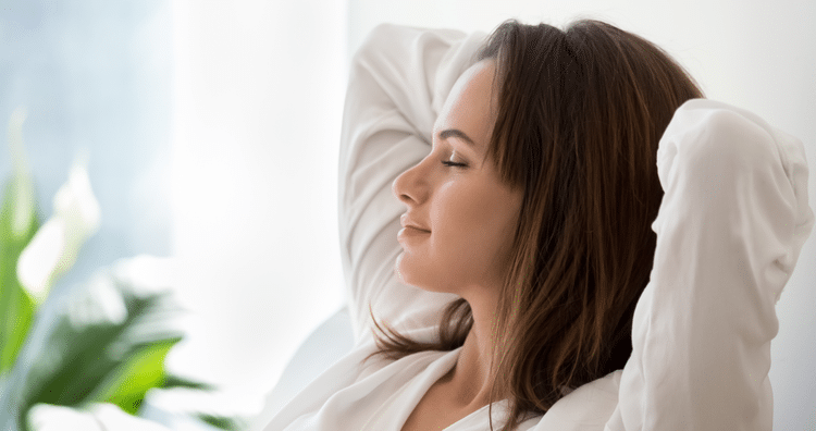 Woman relaxing with her arms behind her head and eyes closed, sitting in a sunny room.