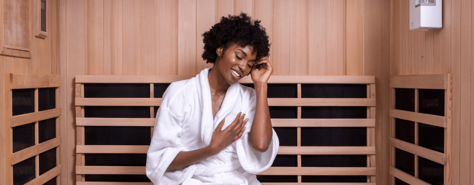 Black woman in a white robe sitting in a sauna relaxing and playing with her hair with eyes closed.