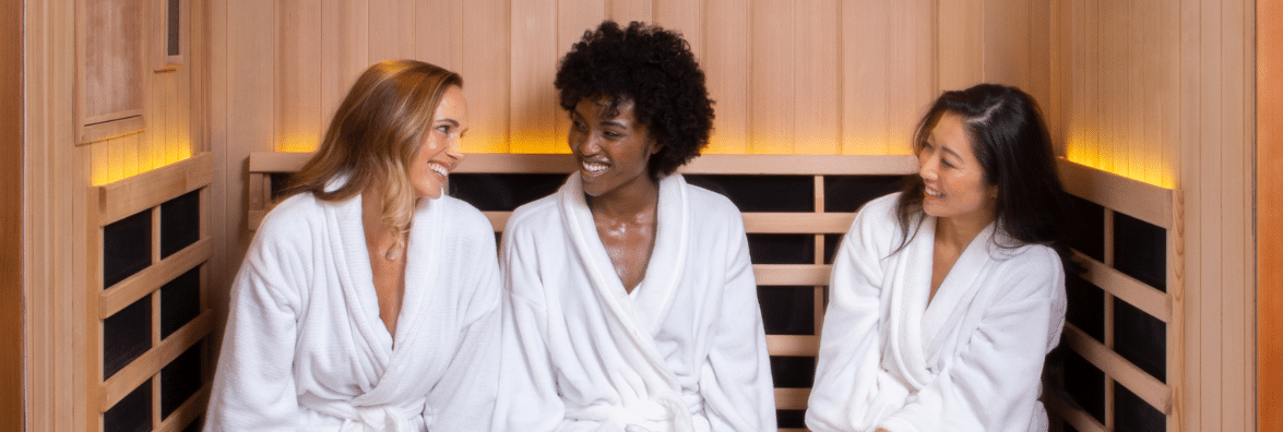 Group of three women in white robes sitting in a sauna talking and laughing.