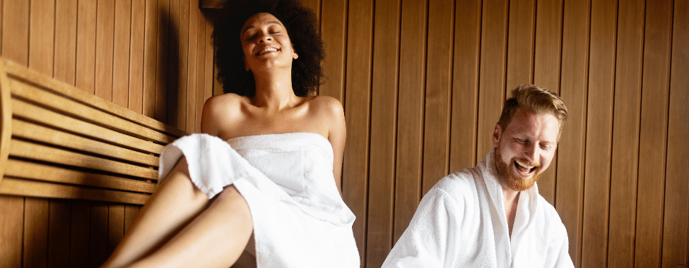 Couple sitting inside a sauna, wearing robes & smiling