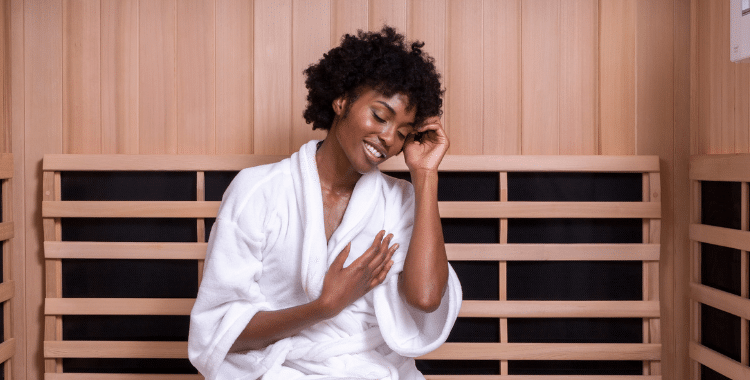 Relaxed woman in a white robe seated in a Clearlight infrared sauna, smiling with her hand on her chest, enjoying the warm ambiance
